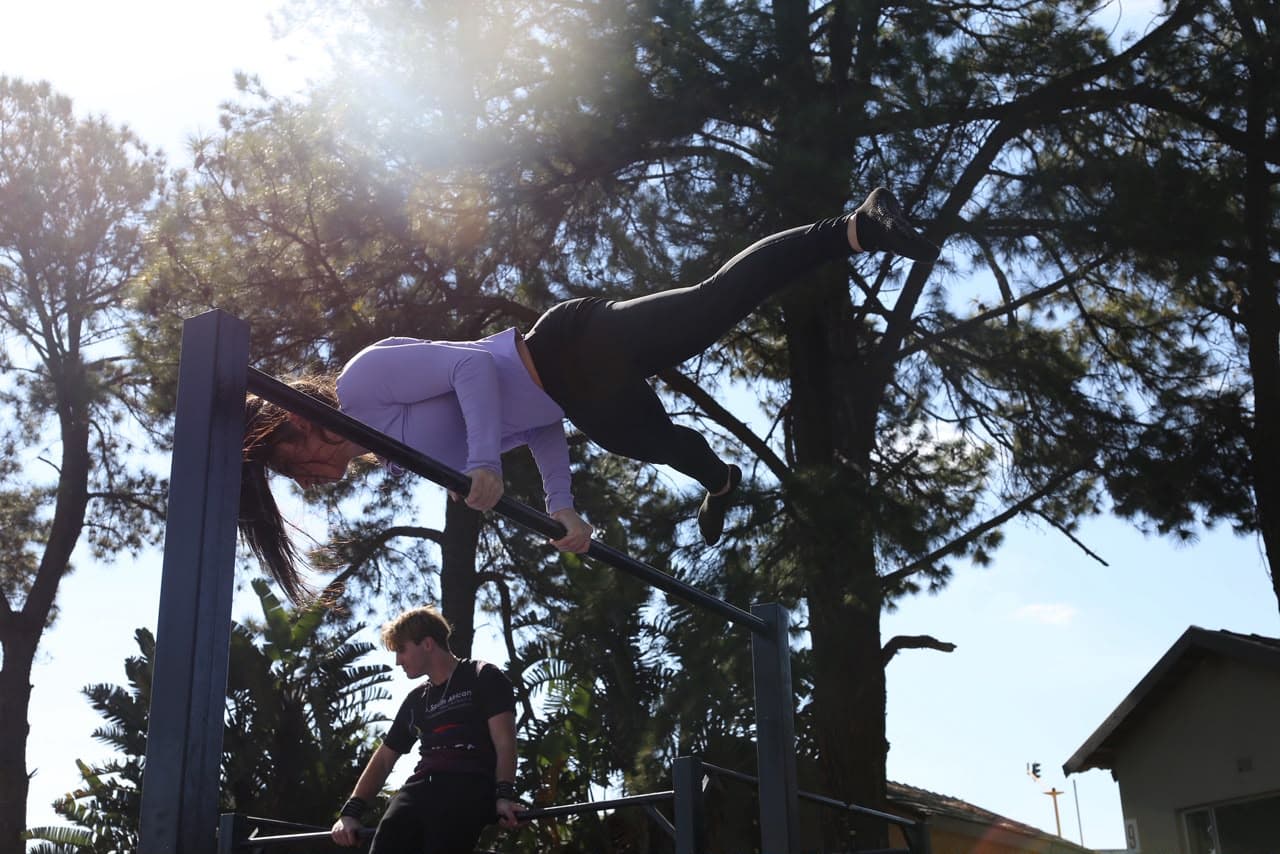 Coach spotting an athlete holding a back lever on the high bar, backlit by low sun