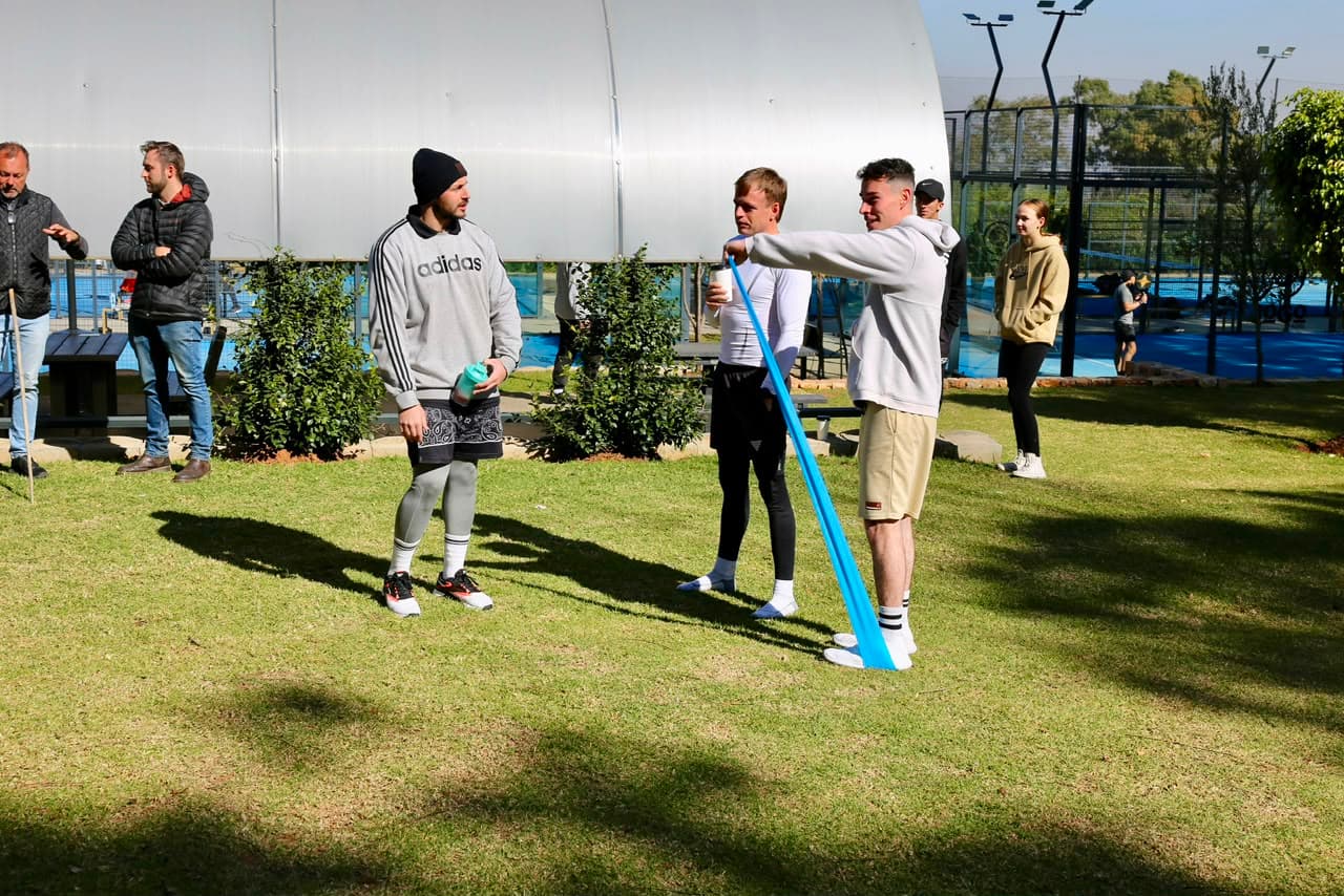 Coach demonstrating a resistance-band exercise to a small group on the grass at the park